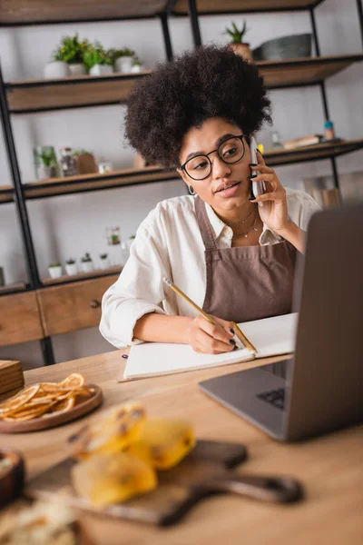 Donna afroamericana in occhiali da vista che parla su smartphone e scrive in notebook vicino a laptop e sapone fatto a mano — Foto stock