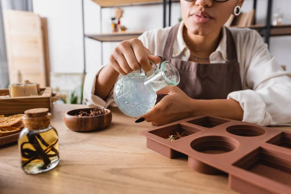 Partial view of african american woman holding jug with liquid soap near silicone mold and natural ingredients in craft workshop — Stock Photo