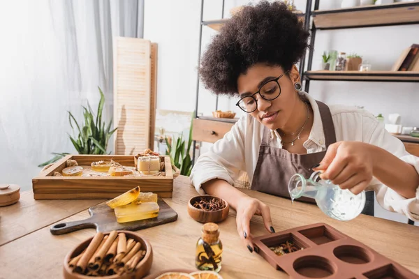 African american woman in apron and eyeglasses pouring liquid soap in silicone mold near spices and essential oils on table - foto de stock