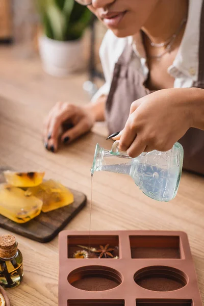 Cropped view of african american woman pouring liquid soap in silicone mold with dried herbs in craft workshop — Stockfoto