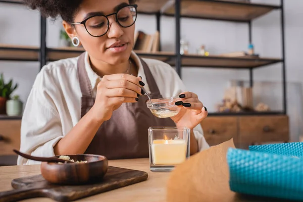 African american craftswoman in eyeglasses heating beeswax above burning candle near wooden bowl and chopping board — Stockfoto