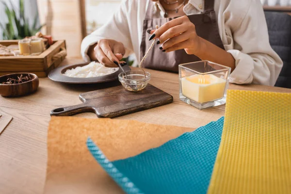 Cropped view of african american craftswoman dipping candle wick in melted beeswax near chopping board and wax sheets - foto de stock