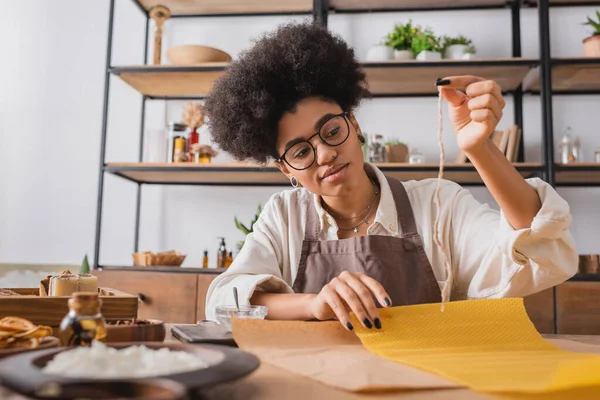 Curly african american woman in eyeglasses and apron holding candle wick near wax sheet and natural ingredients on blurred foreground — Stock Photo
