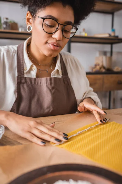 Artisan afro-américain en lunettes et tablier tenant une mèche de bougie près de la feuille de cire sur du papier artisanal — Photo de stock