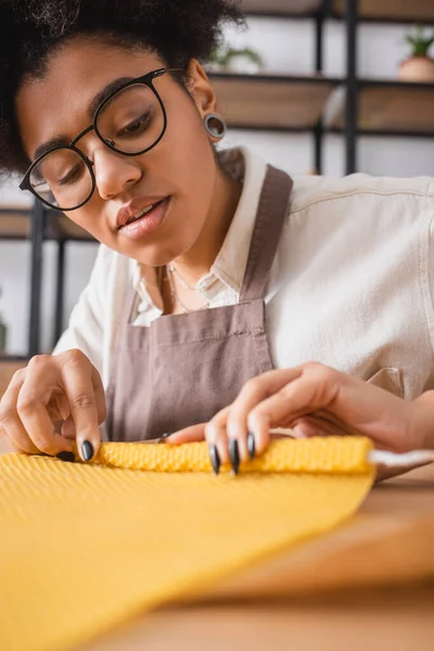 African american woman in apron and eyeglasses rolling wax sheet while making natural candle in craft workshop — Stock Photo