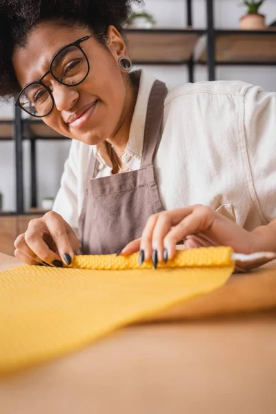 Cheerful african american craftswoman in eyeglasses and apron smiling at camera while making candle in workshop — Stock Photo