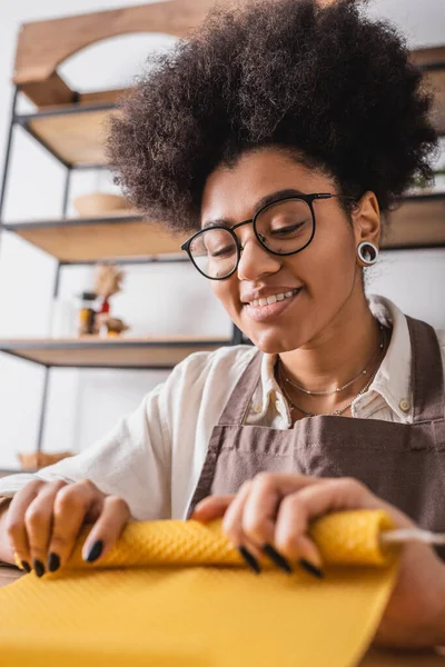 Happy african american woman in eyeglasses and apron rolling natural wax sheet on blurred foreground — Stock Photo