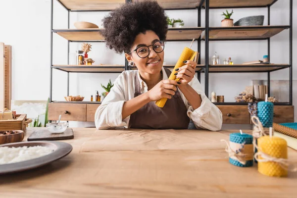 Cheerful african american woman in eyeglasses looking at camera while sitting with handmade candle near parchment in workshop — Stock Photo