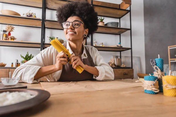 Piacere afro americano donna in occhiali e grembiule tenendo laminato foglio di cera vicino a candele fatte in casa in laboratorio artigianale — Foto stock