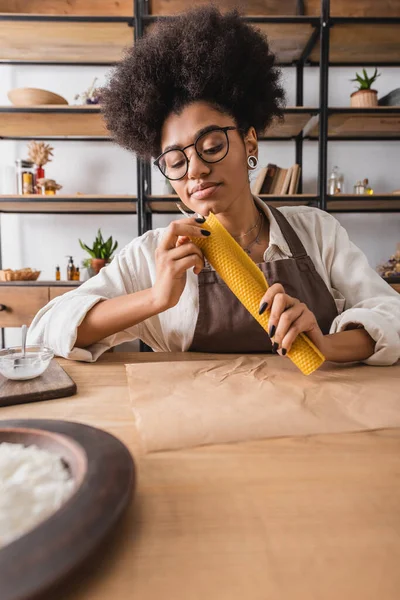 Artesana afroamericana rizada en delantal y anteojos haciendo mango natural cerca de pergamino en mesa en taller - foto de stock