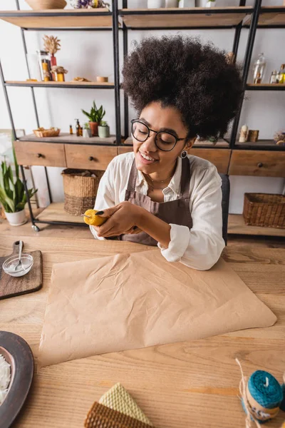 Sorridente artigiana afroamericana in grembiule e occhiali che fa candela naturale vicino alla carta artigianale in officina — Foto stock