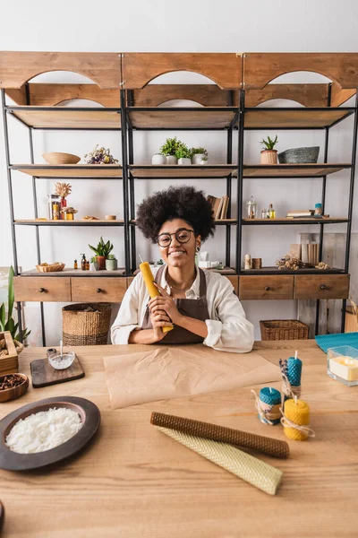 Heureuse femme afro-américaine tenant une feuille de cire roulée près du parchemin et des bougies faites à la main sur la table dans un atelier artisanal — Photo de stock