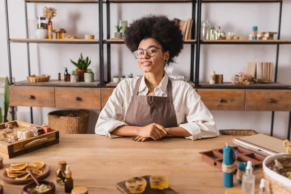 Felice artigiana afroamericana in occhiali e grembiule guardando la fotocamera vicino a prodotti naturali e ingredienti in officina — Foto stock