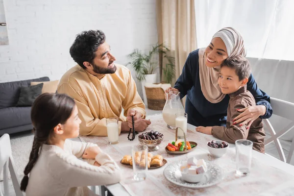 Smiling african american woman in hijab hugging son near family during suhur breakfast in morning — Foto stock