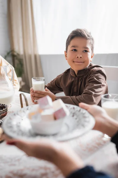 Muslim boy holding glass of milk near mother holding blurred turkish delight during ramadan in morning — Stock Photo