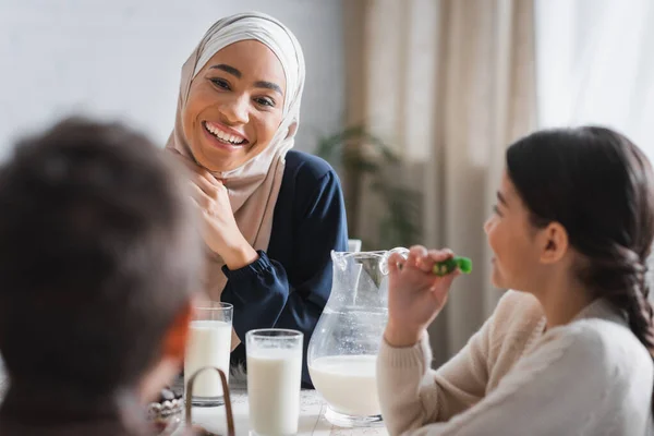 Sonriente madre afroamericana en hijab mirando a los niños borrosos cerca de la comida de suhur en casa - foto de stock