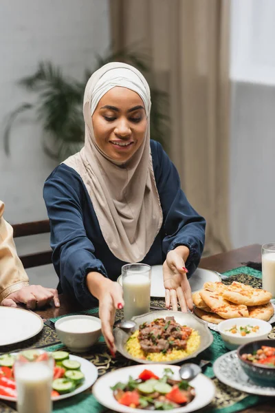 Positive african american woman in hijab putting food on table during ramadan dinner at home - foto de stock
