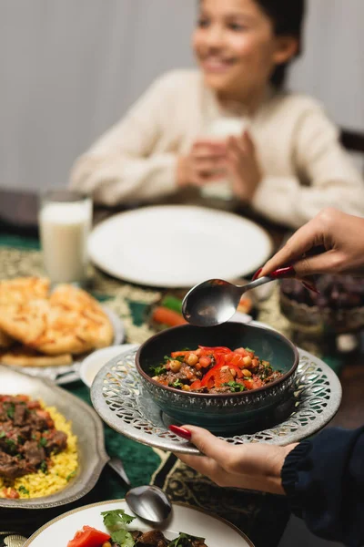 Muslim woman holding tasty dish near blurred daughter and ramadan dinner — Stock Photo