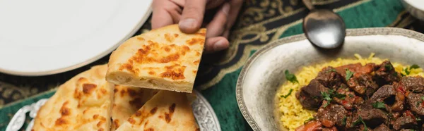 Cropped view of muslim man holding tasty pita bread near ramadan dinner at home, banner - foto de stock