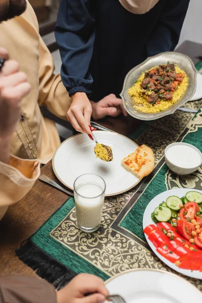 Cropped view of muslim woman serving pilaf on plate near family during ramadan — Stockfoto
