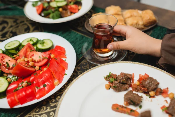 Vista ritagliata di ragazzo musulmano che tiene tradizionale tazza di tè di vetro vicino al cibo a casa — Foto stock