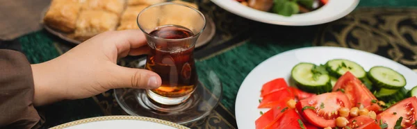 Vue recadrée du garçon musulman tenant une tasse de thé traditionnelle en verre près de la nourriture iftar à la maison, bannière — Photo de stock