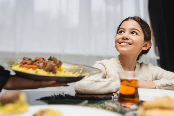 Smiling muslim girl looking at blurred mom with pilaf at home — Photo de stock