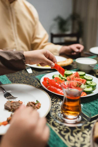 Vista ritagliata del ragazzo musulmano che prende verdura fresca vicino al padre offuscata e cena al ramadan — Foto stock
