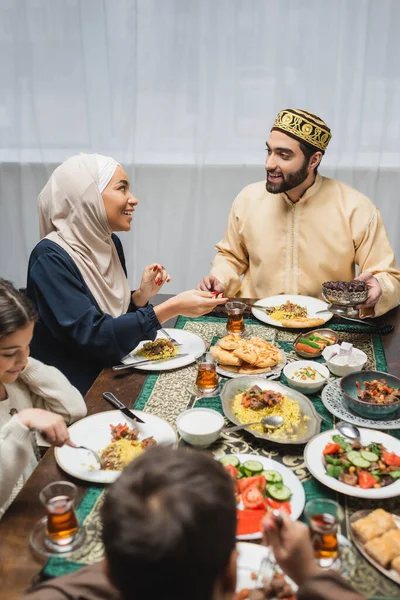Sonrientes padres musulmanes hablando cerca de los niños y la cena iftar en casa - foto de stock