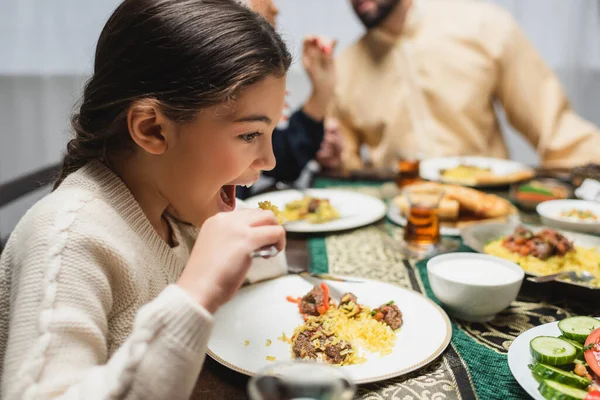Muslim girl eating pilaf near blurred parents and ramadan dinner — Stock Photo