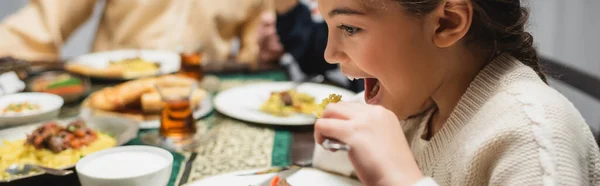 Muslim girl eating iftar dinner near blurred food and parents at home, banner — Stockfoto