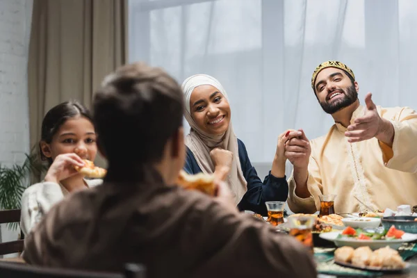 Sorridenti genitori musulmani che si tengono per mano vicino a bambini sfocati e cibo durante il ramadan a casa — Foto stock