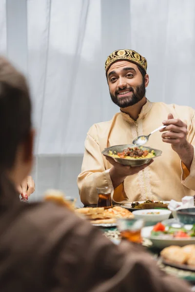 Padre musulmán positivo sosteniendo comida cerca de la familia borrosa durante el ramadán en casa - foto de stock