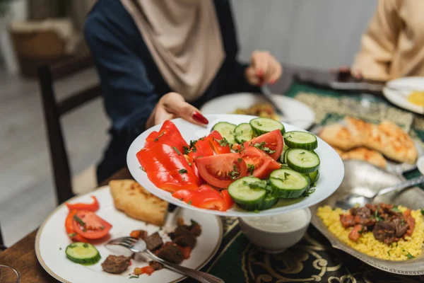 Cropped view of muslim woman holding plate with vegetables near food during iftar at home — Stockfoto