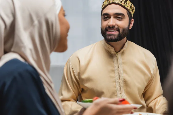 Positive muslim man looking at blurred wife with food at home — Stock Photo