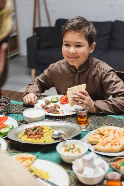 Muchacho musulmán sonriente sosteniendo pan de pita cerca de papá borroso y comida en casa - foto de stock