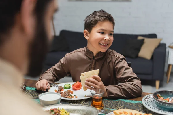 Cheerful muslim boy looking away near food and dad during iftar at home — Stockfoto