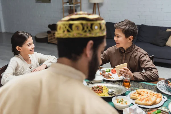 Niños musulmanes positivos sentados cerca de la comida y padre borroso durante el ramadán en casa - foto de stock