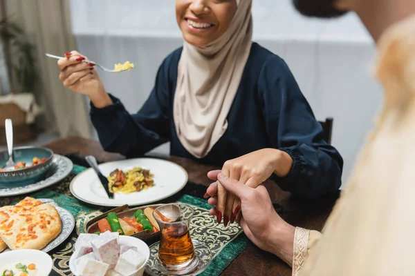 Vista recortada de pareja musulmana cogida de la mano cerca de la comida durante iftar en casa - foto de stock