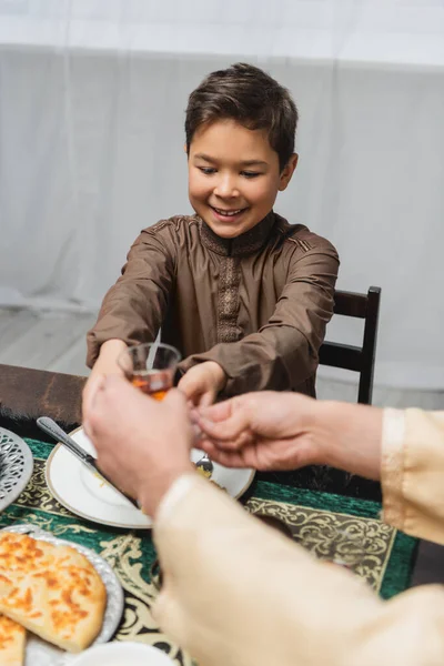 Smiling muslim boy giving turkish tea glass to blurred dad during iftar at home — Foto stock