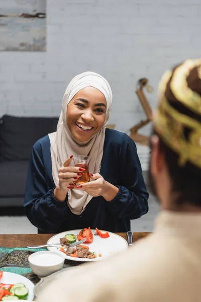 Femme afro-américaine souriante dans le hijab tenant le thé près du mari flou pendant iftar à la maison — Photo de stock