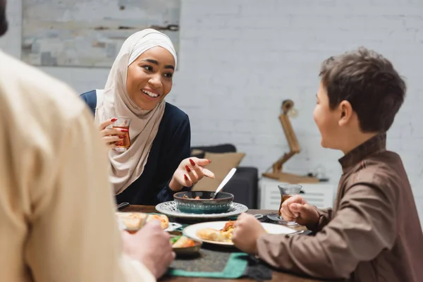 African american woman in hijab holding tea and talking to son near husband and food at home — Stockfoto