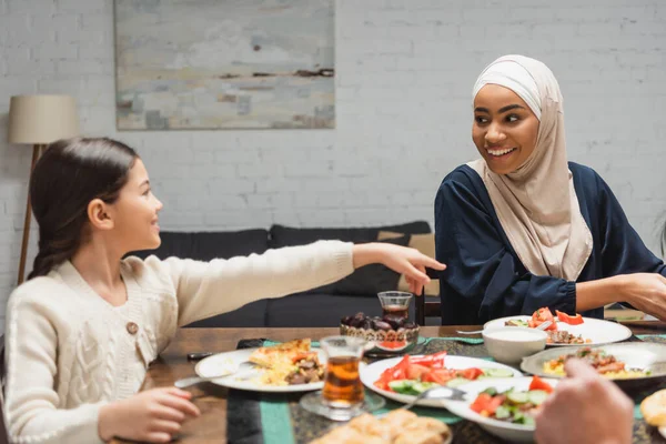 Sonriente madre afroamericana en hijab mirando a su hija cerca de la comida iftar en casa - foto de stock