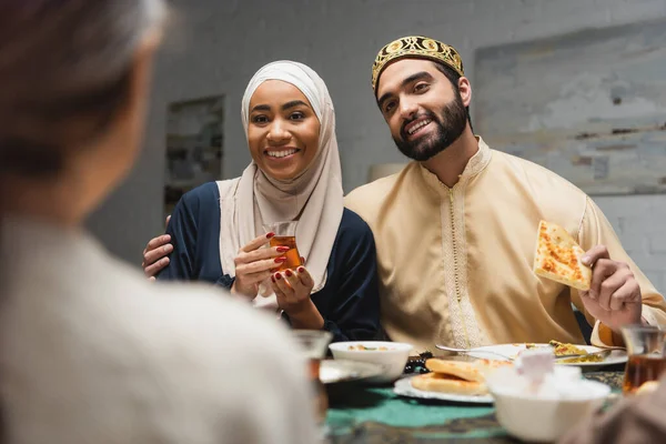 Homem muçulmano abraçando esposa no hijab perto de filha turva e comida durante o ramadã em casa — Fotografia de Stock