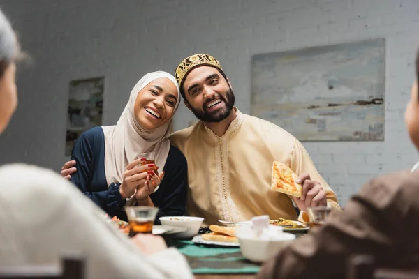 Smiling muslim man hugging wife with tea near blurred kids and food at home — Stockfoto