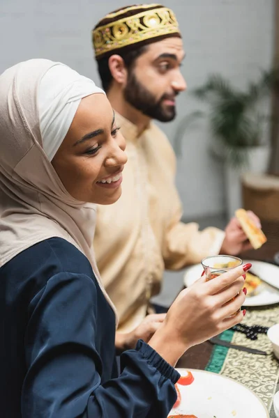 Smiling african american woman in hijab holding tea glass near blurred husband during ramadan at home - foto de stock