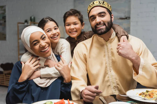 Niños sonrientes abrazando a padres musulmanes y mirando a la cámara durante iftar en casa - foto de stock