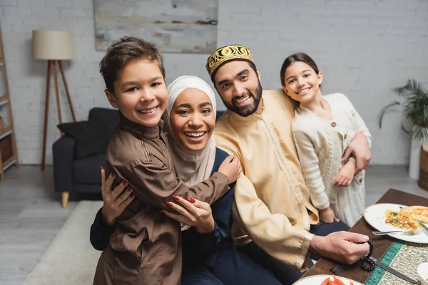 Positive middle east family looking at camera near dinner during ramadan at home - foto de stock