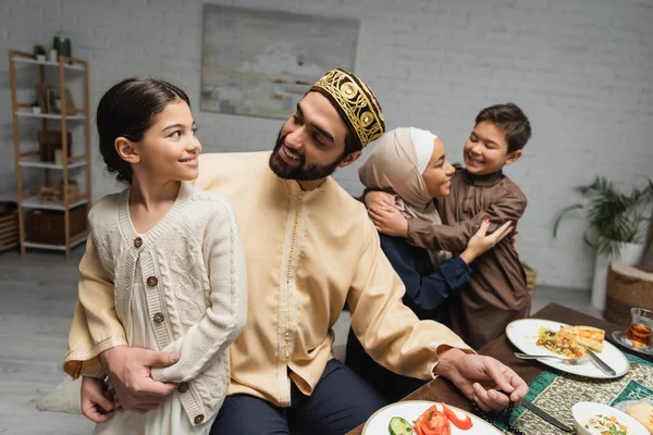 Padre musulmán abrazando a su hija cerca de la familia y la cena de ramadán en casa - foto de stock