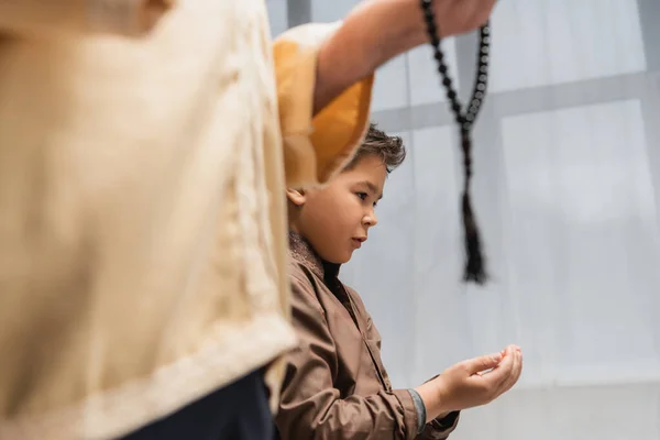 Middle eastern boy praying near blurred dad with prayer beads at home — Stock Photo